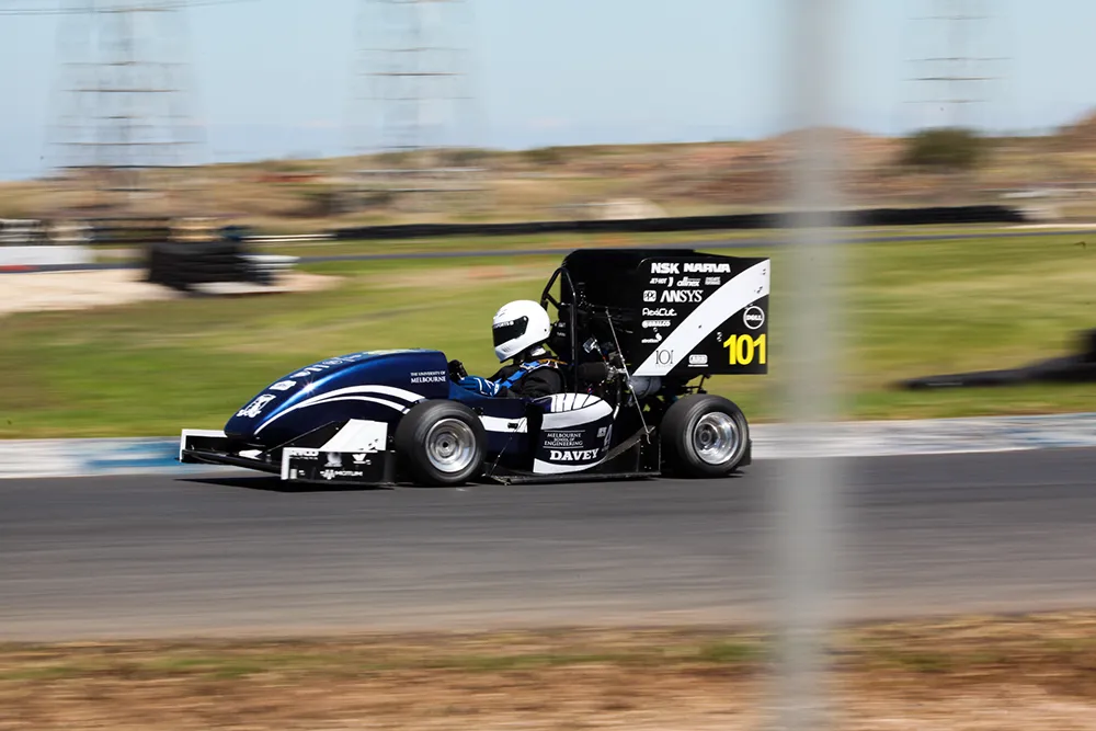 Two professional women talking in an office; one holds a tablet while the other gestures, with colleagues working in the background.